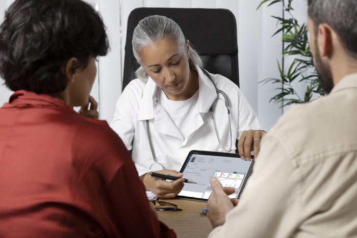 A doctor sitting at a desk with two patients, discussing medical information displayed on a tablet.