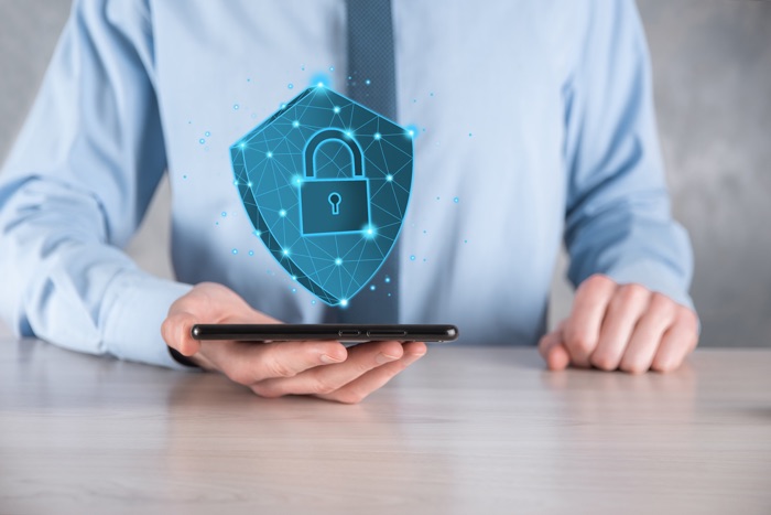 A person in business attire holding a smartphone with a holographic image of a lock and shield above it, symbolizing security.